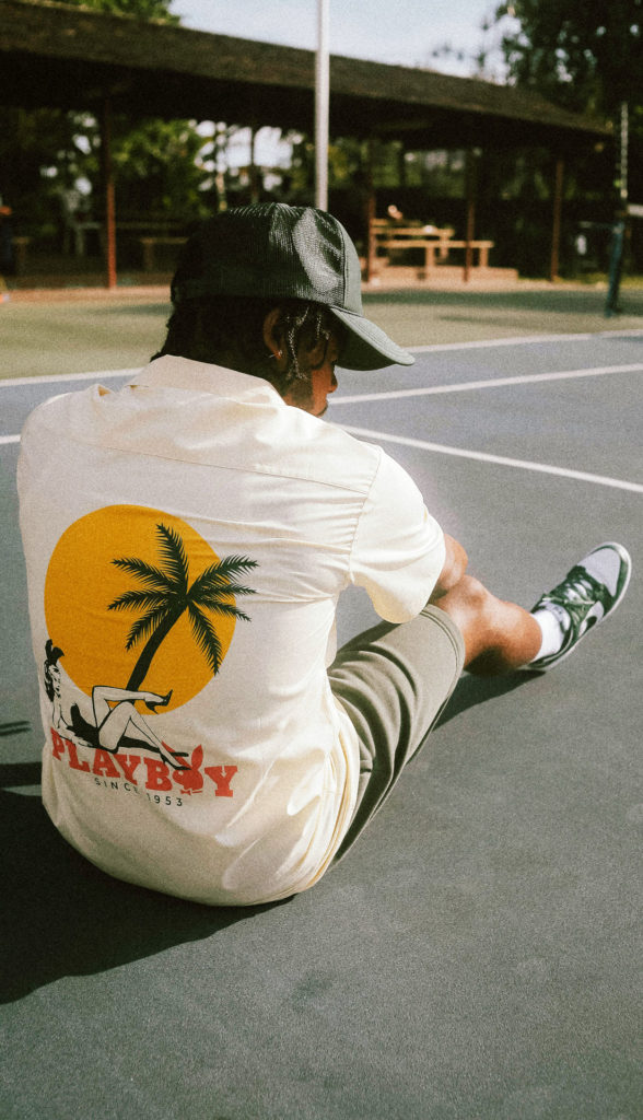 Person sitting on a tennis court in a casual outfit with a tropical-themed shirt, cap, and trainers.