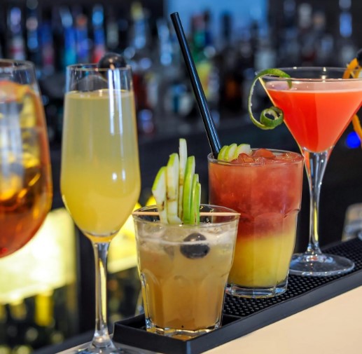 Assorted cocktails on a bar counter, featuring vibrant colours and garnishes, set in a lively, well-lit pub setting.