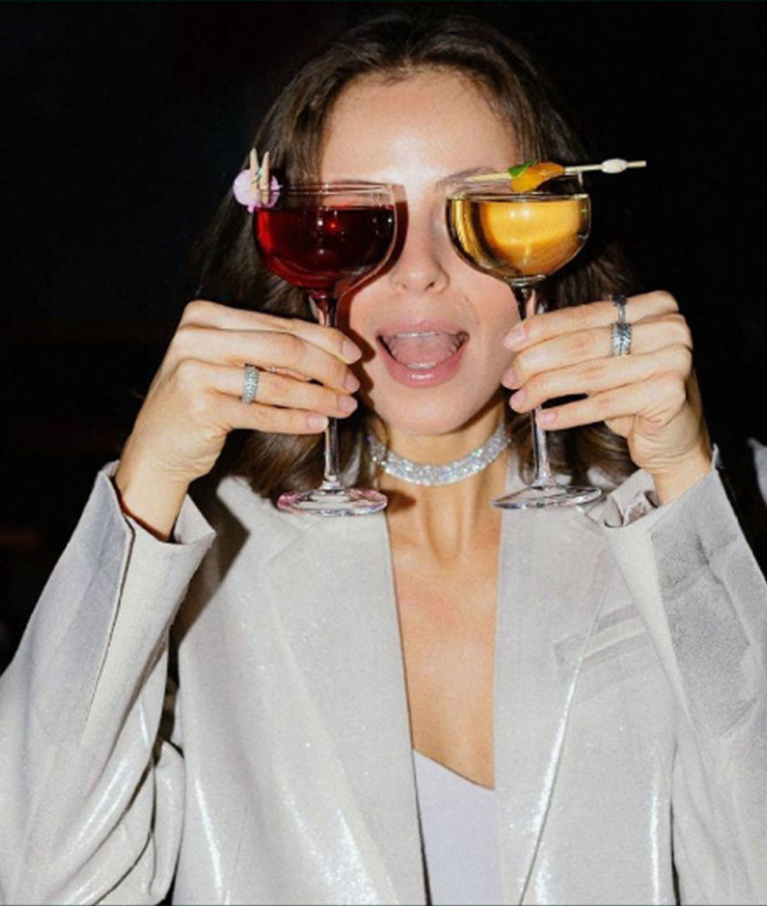 Woman playfully holding two cocktails like glasses. Red and orange drinks add a fun touch to her shimmering outfit.