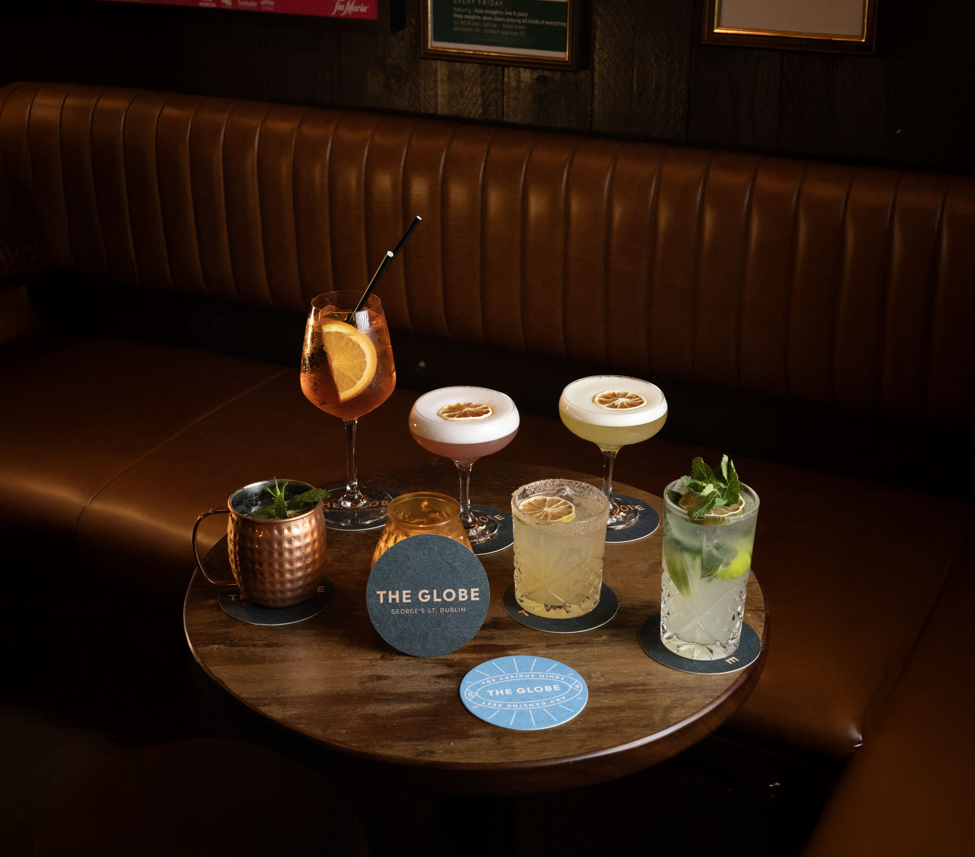 A wooden table in a cozy bar setting holding six different cocktails, including a spritz with an orange slice, two foam-topped drinks with dried citrus garnishes, a Moscow mule in a copper mug, a lime-garnished highball, and a minty cocktail, with round coasters including one that says ‘The Globe’.